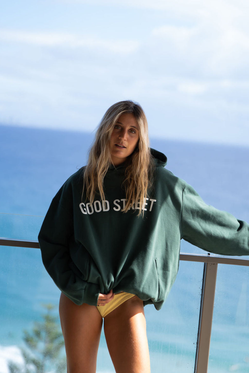 Person wearing a green 'GOOD STREET' hoodie standing by a railing with a scenic background.